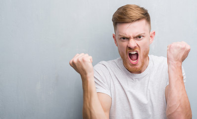 Young redhead man over grey grunge wall annoyed and frustrated shouting with anger, crazy and yelling with raised hand, anger concept