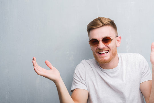 Young redhead man over grey grunge wall wearing retro sunglasses very happy and excited, winner expression celebrating victory screaming with big smile and raised hands