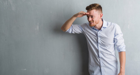 Young redhead business man over grey grunge wall very happy and smiling looking far away with hand over head. Searching concept.