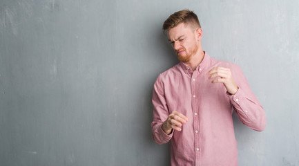 Young redhead man over grey grunge wall wearing pink shirt disgusted expression, displeased and fearful doing disgust face because aversion reaction. With hands raised. Annoying concept.