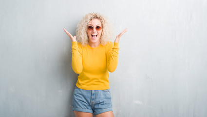 Young blonde woman with curly hair over grunge grey background celebrating crazy and amazed for success with arms raised and open eyes screaming excited. Winner concept