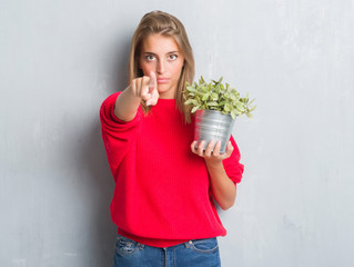 Beautiful young woman over grunge grey wall holding plant pot pointing with finger to the camera and to you, hand sign, positive and confident gesture from the front
