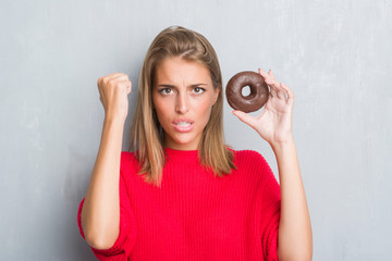 Beautiful young woman over grunge grey wall eating chocolate donut annoyed and frustrated shouting with anger, crazy and yelling with raised hand, anger concept