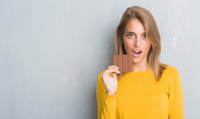 Beautiful young woman over grunge grey wall eating chocolate bar with a confident expression on smart face thinking serious