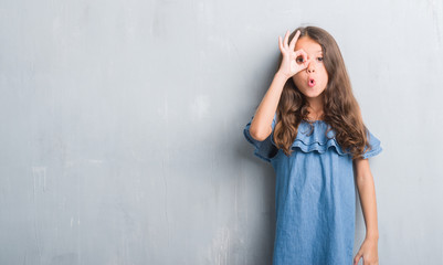 Naklejka premium Young hispanic kid over grunge grey wall doing ok gesture shocked with surprised face, eye looking through fingers. Unbelieving expression.