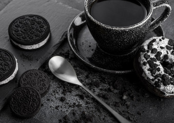 Obraz na płótnie Canvas Black coffee cup with saucer and doughnut with black sandwich cookies on black stone kitchen table background. Space for text. Breakfast snack. Black cookie ice cream on stone board with silver spoon