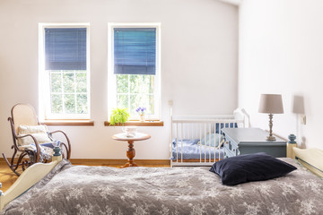 A nursery room interior with a bed for adults, a white cot for a child an a rocking chair. Windows...