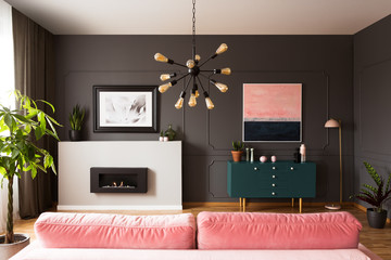 Lamp above pink couch in grey apartment interior with green cabinet and fireplace. Real photo