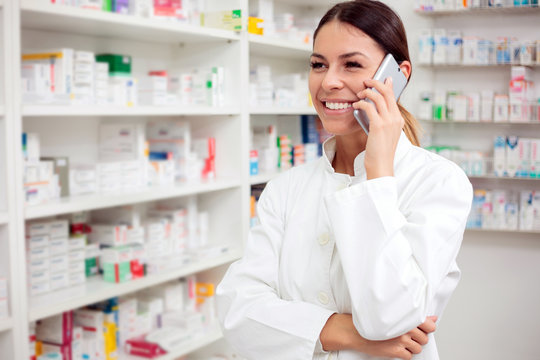 Beautiful young female pharmacist talking on the phone, shelves with medications in the background