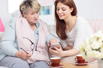 Smiling daughter showing smartphone to senior woman while drinking tea
