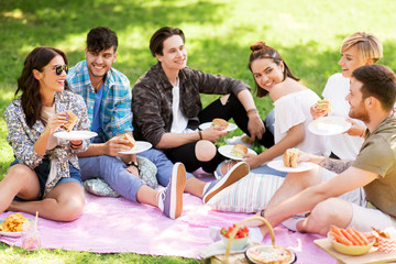 friendship, leisure and fast food concept - group of happy friends eating sandwiches or burgers at picnic in summer park