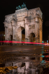 Fototapeta na wymiar The Siegestor Victory Arch in Munich. Triumphal arch at night on a rainy day. Side view with a puddle and reflections in the foreground.