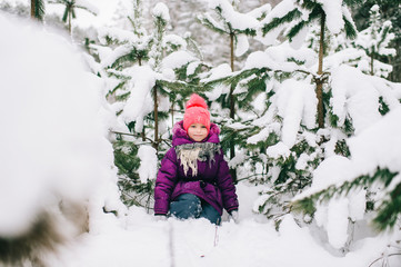 Adorable little girl posing under snowy spruce tree in wood outdoor. Beautiful female child sitting on snow in front of christmas tree in forest. Winter portrait of lovely young baby.  Funny kid face.