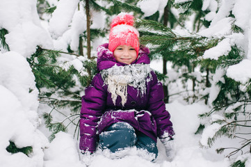 Adorable little girl posing under snowy spruce tree in wood outdoor. Beautiful female child sitting on snow in front of christmas tree in forest. Winter portrait of lovely young baby.  Funny kid face.