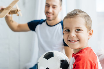 selective focus of smiling boy with football ball and father with wooden toy plane at home