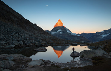 Hiker on a rock in the Riffelsee watching the first sunlight  on the Matterhorn. - obrazy, fototapety, plakaty