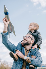happy father carrying little son on neck and holing model rocket against sky
