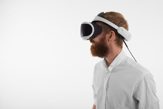 Visual reality and artificial intelligence concept. Profile shot of bearded red haired young man wearing oculus rift headset against blank studio wall background with copy space for your text