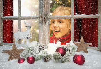 Lachendes Kind schaut freudig überrascht aus dem weihnachtlich dekorierten Weihnachtsfenster,...