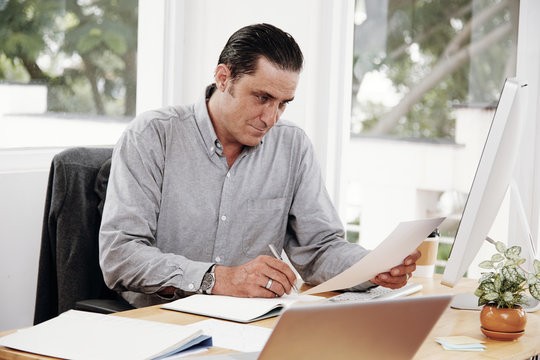 Entrepreneur writing notes sitting at his desk. Serious man making business plans with papers and computers on his desk