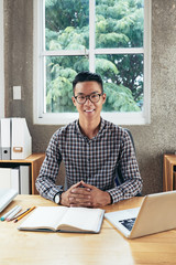 Portrait of young attractive confident Asian man in glasses sitting at table with notebook and smiling at camera happily