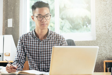 Young concentrated handsome Asian man sitting at desk, reading information on laptop and taking notes in notepad
