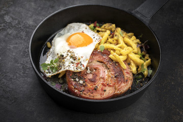 Traditionelle gebratene Allgäuer Kalbsrolle  aufgeschnitten mit Spiegelei und Spätzle in einer gusseisernen Pfanne mit Gewürzen als closeup auf einem schwarzen Board
