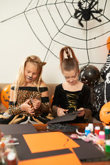 Pretty Caucasian girls in smart costumes with painted faces cutting out colored paper while preparing for Halloween celebration