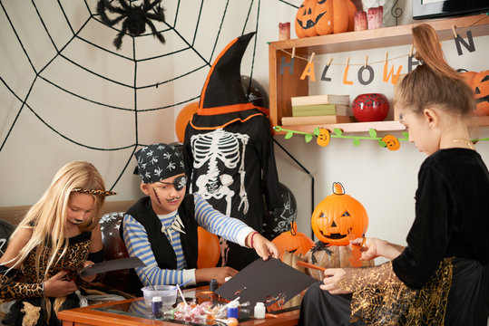 Group of little Caucasian boys and girls in Halloween costumes making diy paper decorations while sitting in beautifully decorated room