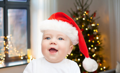 holidays, children and people concept - close up of happy little baby boy or girl in santa hat at home over christmas tree lights background