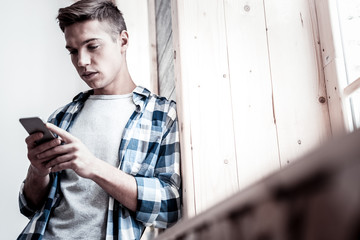 By the window. Calm young man wearing casual clothes at home and thoughtfully looking at the screen of his device while standing by the window