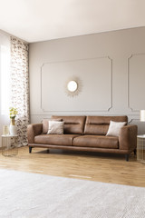 Pillows on leather sofa in elegant grey living room interior with flowers and gold mirror. Real photo