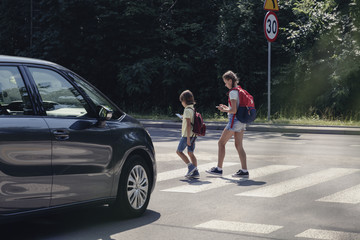 Car in front of children on pedestrian crossing walking from the school and looking at their smartphones - obrazy, fototapety, plakaty