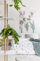 Close to nature bright bedroom interior with a bed covered with white linen and marine blanket. Green plants on shelves next to the bed. Fabric art wall. Real photo.