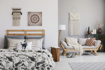 Poster and decor above patterned bed in hygge flat interior with pillows on wooden sofa. Real photo