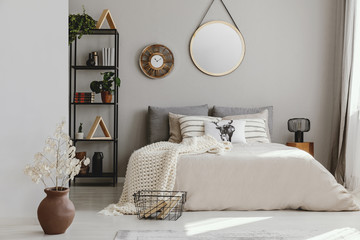 Round mirror and clock above bed with pillows in bright bedroom interior with flowers. Real photo