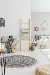 Pouf and round rug in bright living room interior with ladder next to wooden couch. Real photo