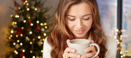 holidays and people concept - happy young woman with cup of coffee or tea at home over christmas tree lights background