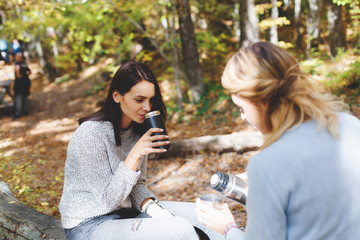 Two young girls girlfriend sat down to rest and drink tea from thermo cup in the autumn forest. ...