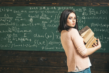Course project. Girl holds heavy pile of old books, chalkboard background. Girl student works on...