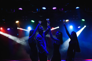 Fototapeta premium Young friends dancing with glasses of champagne in hands. Against lighting devices as background. Young people's friends are dancing.