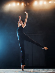 Young beautiful ballerina on smoke stage dancing modern ballet. performs smooth movements with hands against spotlights background. Woman in black costume on scene