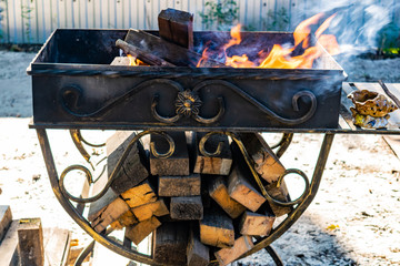 Fototapeta na wymiar Metal grill, forged, handmade, with a pattern. A hot fire on the wood with smoke for barbecue and grilled products. Outside on a Sunny day.