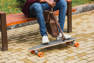 cropped shot of man with leather backpack and skateboard sitting on bench on street