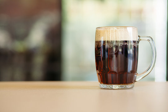 Horizontal photo of glass cup full of beer standing on smooth wooden surface. Cold dark summer drink for day heat. Fresh with thick high foam. Light blurred background. Concept of drinks shooting.