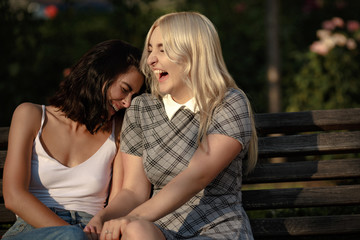 Fototapeta premium Two beautiful young girls sitting on bench and laughing. Holding hands. Lesbian love concept.