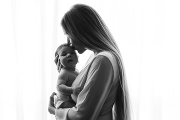 black and white picture of mother kissing little baby boy in front of curtains at home