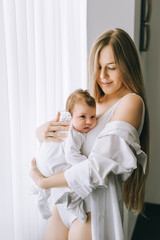 smiling mother carrying little baby boy in front of curtains at home