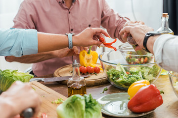 cropped image of mature friends preparing salad for dinner at home and putting ingredients in bowl