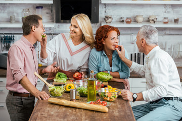 happy mature couples feeding each other during salad preparation for dinner at home